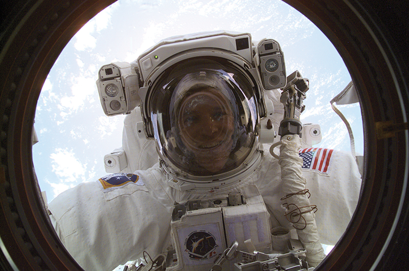 Astronaut Scott Parazynski in a spacesuit looks through a window on the space station with Earth behind him