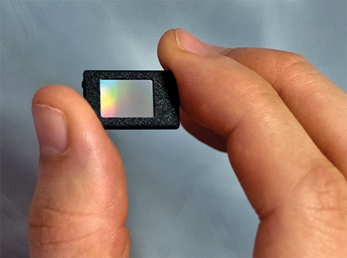 A thumb and forefinger hold a thumbnail-sized microdisplay panel.