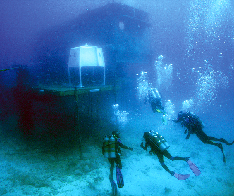 Crew members in scuba gear swim toward the underwater NEEMO laboratory