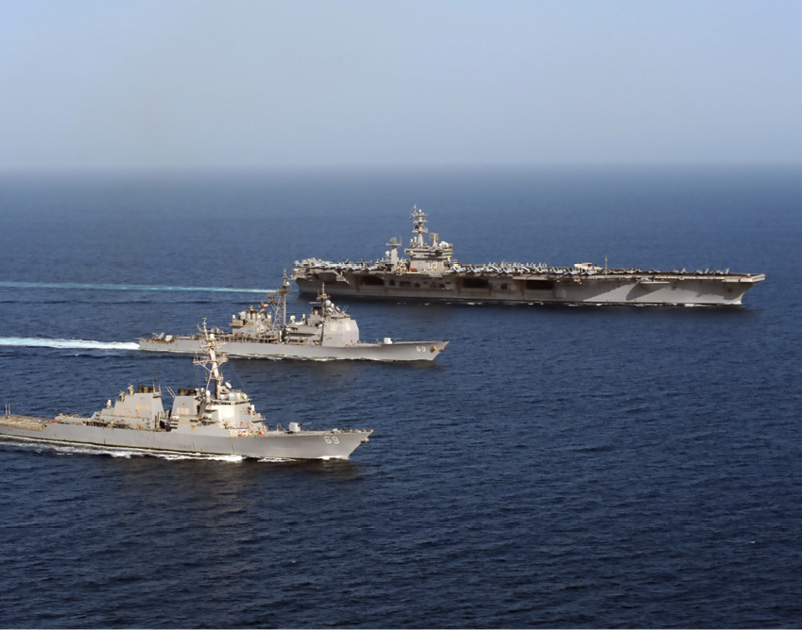 A U.S. Navy aircraft carrier and two more ships navigate in parallel on deep blue water