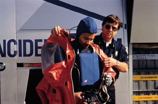 Hazmat team member is helped into a protective vapor suit by a teammate. The chest pack includes a cooling cartridge and controls