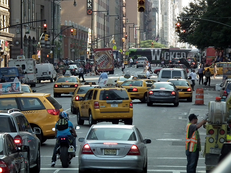 A view of a crowded New York City street, with pedestrians, bikes, buses, trucks, cars, and yellow cabs