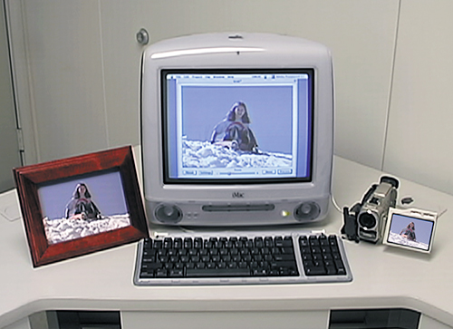 An imac with an image on the screen, a framed image and a video camera beside it.