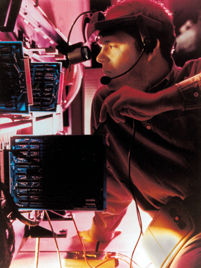 A man wearing a headset looks through a scope at a rack of circuit boards