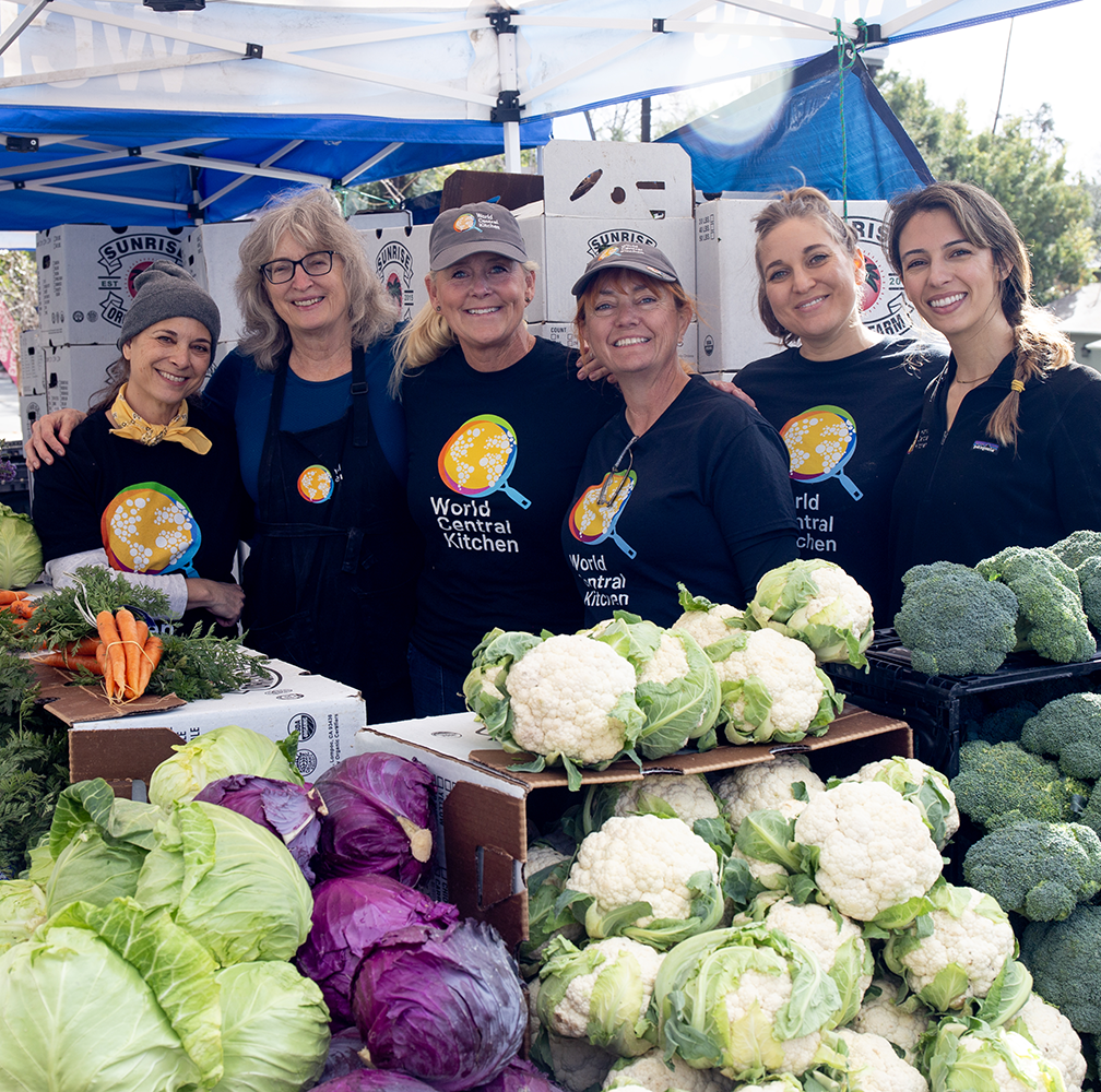 Women wearing World Central Kitchen T-shirts stand behind an assortment of vegetables 