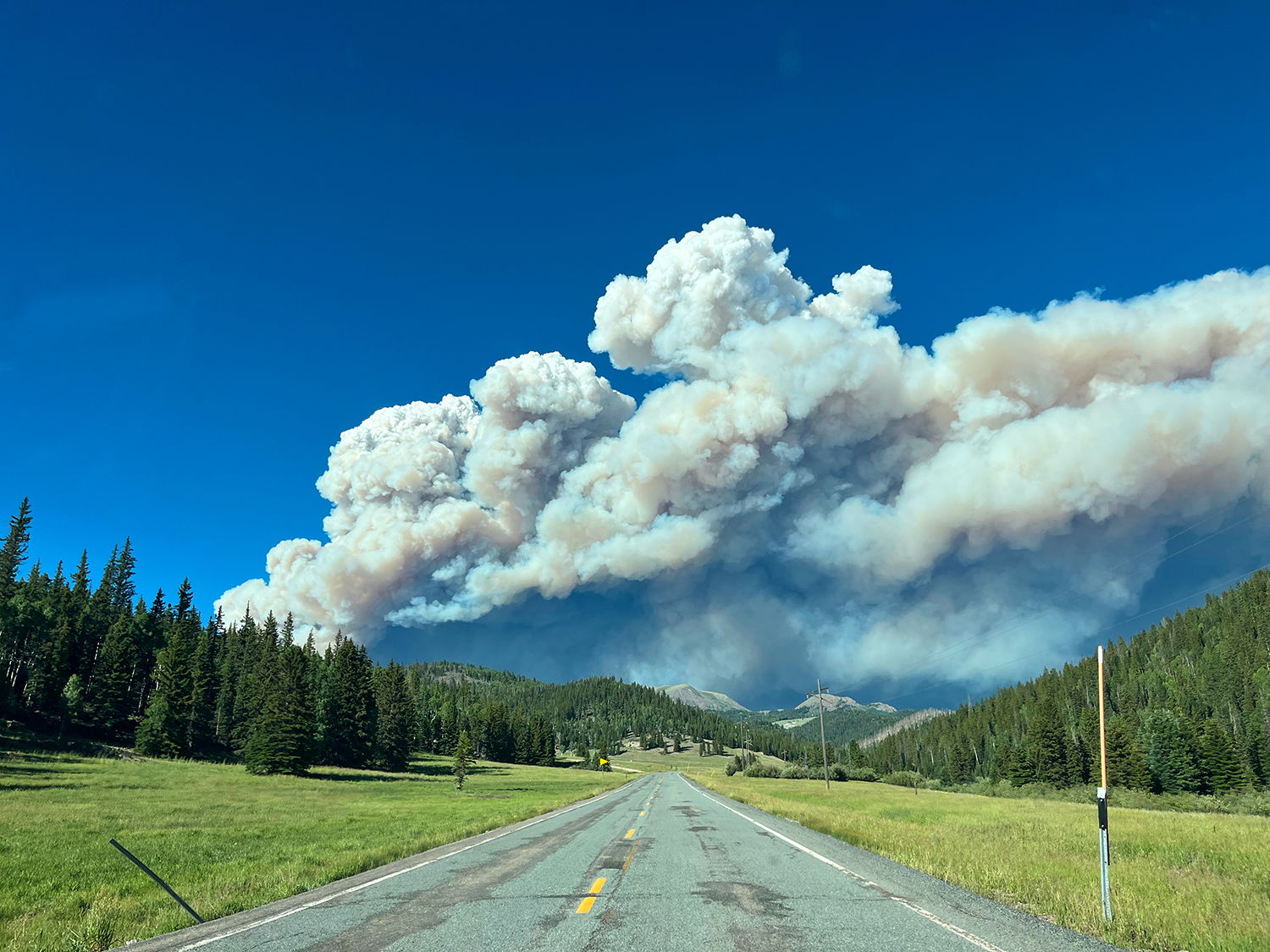 road through a landscape of grass, pines, and hills with billowing smoke above
