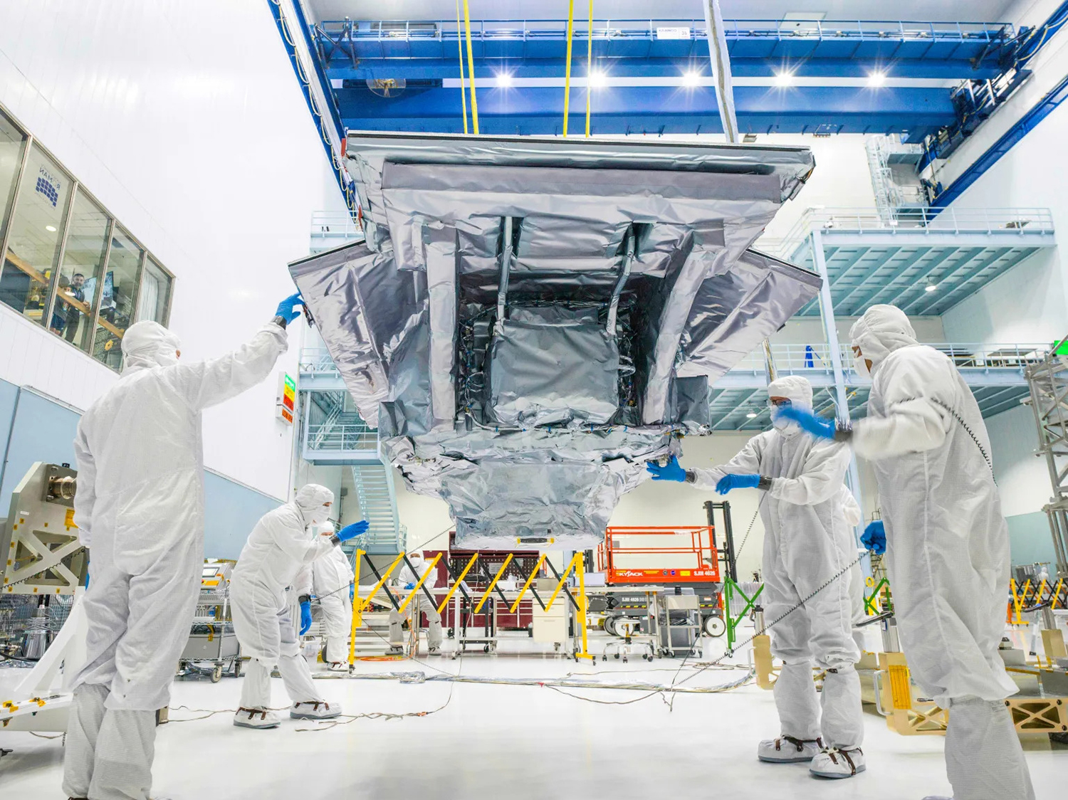 Four technicians in white protective suits working on a large suspended instrument 