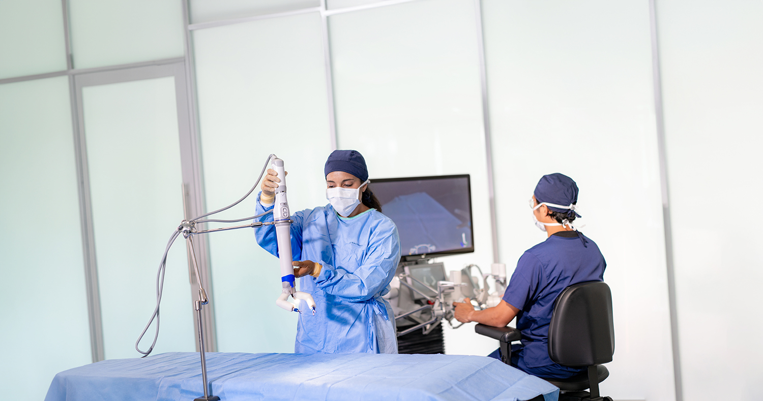 Man in scrubs sitting at a computer behind a woman in scrubs positioning device over operating table