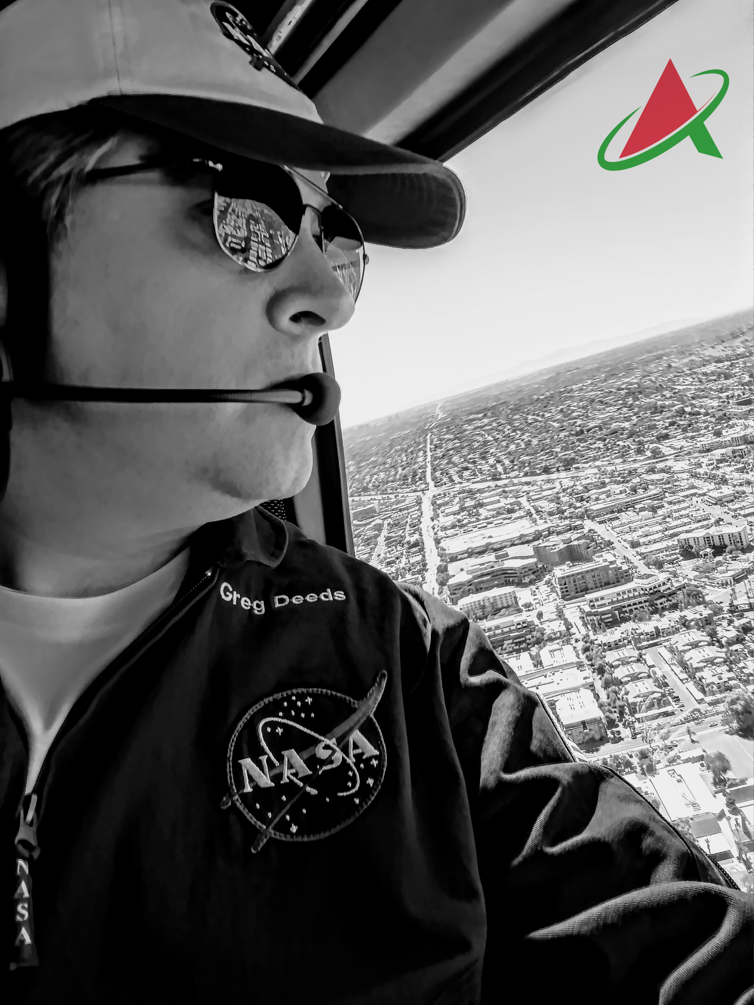 A black and white photo of a man wearing a shirt emblazoned with the NASA emblem looking out of the window of a helicopter at the city below.