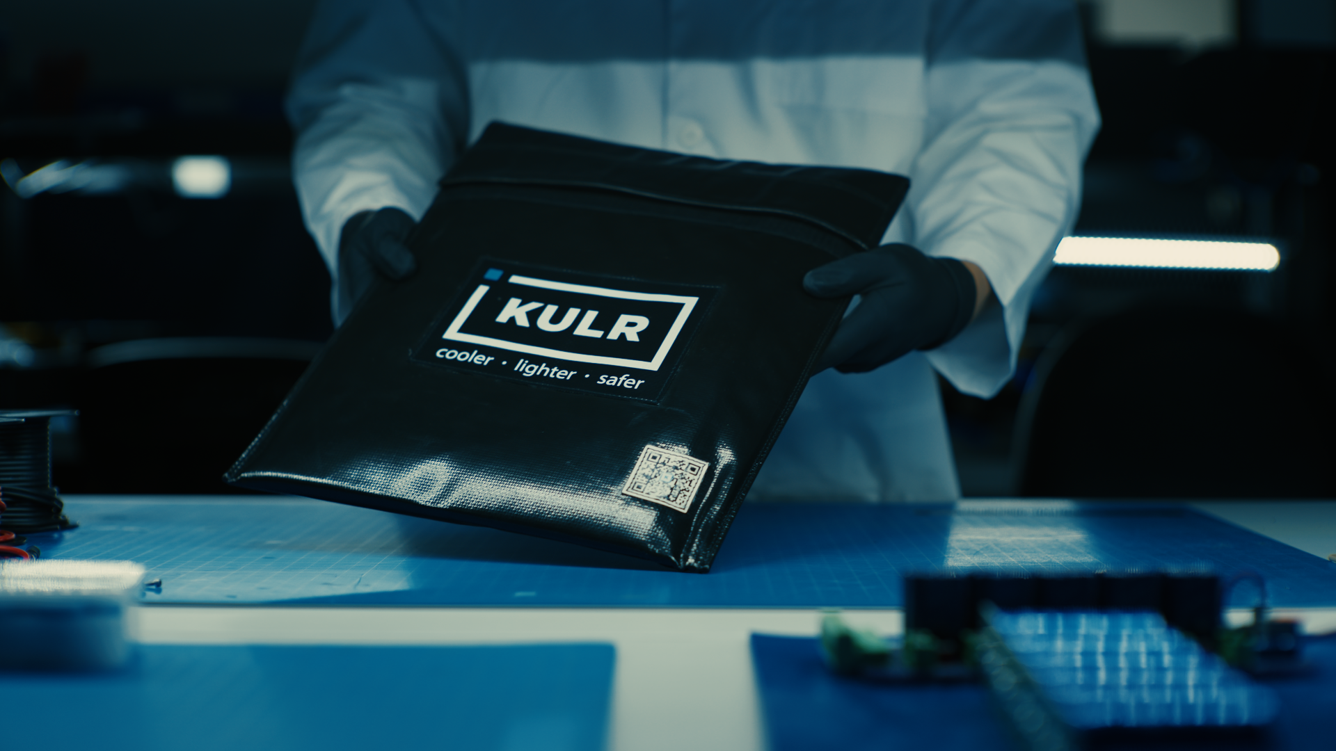 A person in a lab coat and gloves holding a black pouch. Text on the pouch reads “KULR: cooler • lighter • safer.”