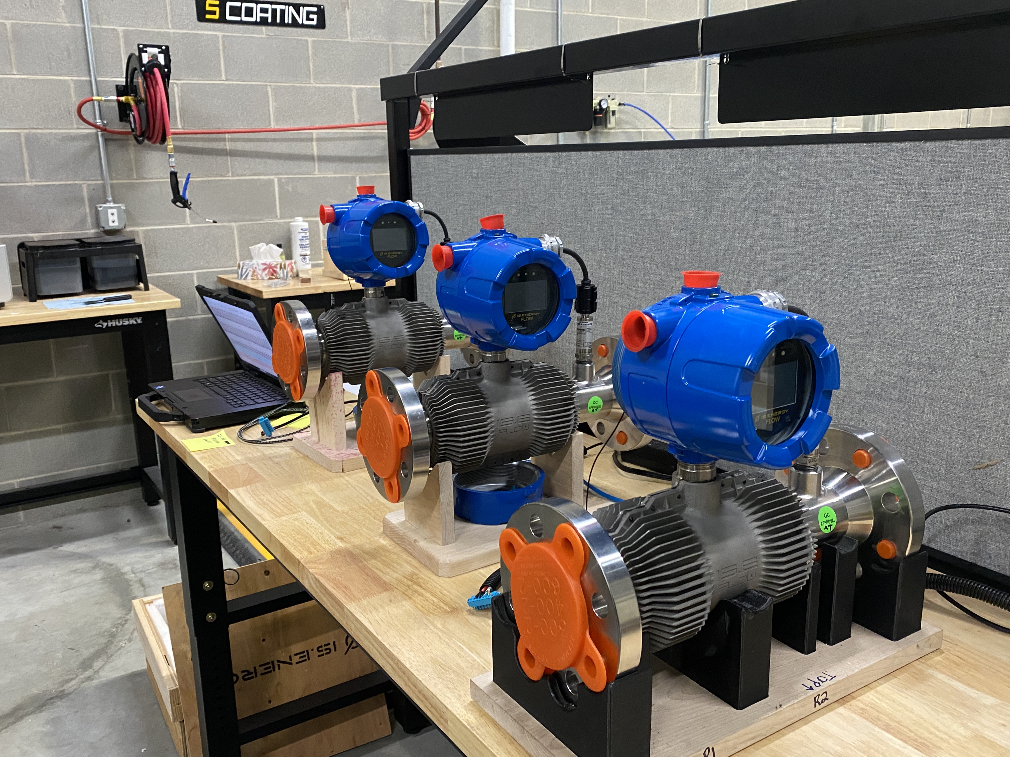 On a wooden table are three blue and orange machines next to a laptop computer