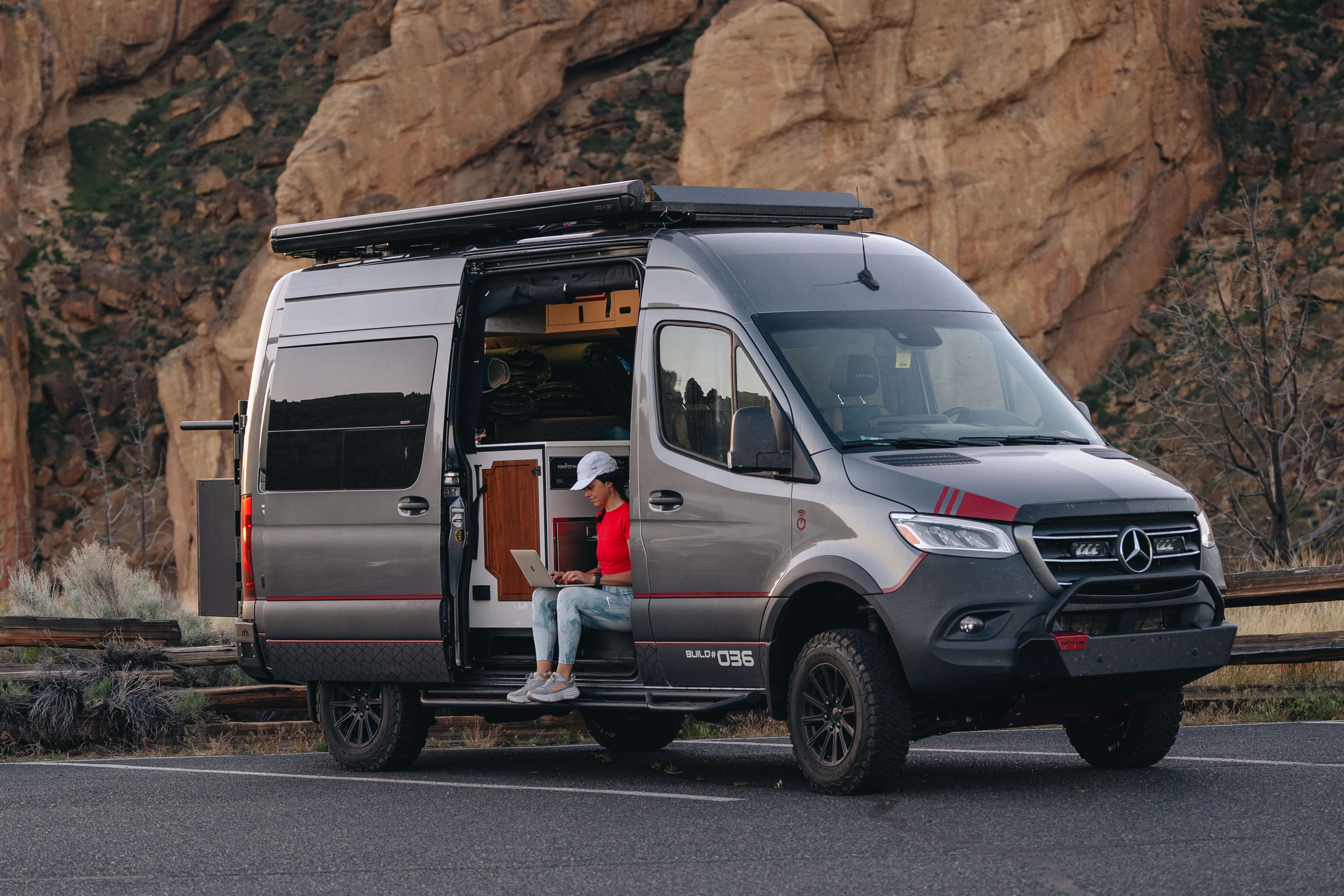 On the side of a mountain road, A woman works on a laptop while sitting in the open side door of a camper van