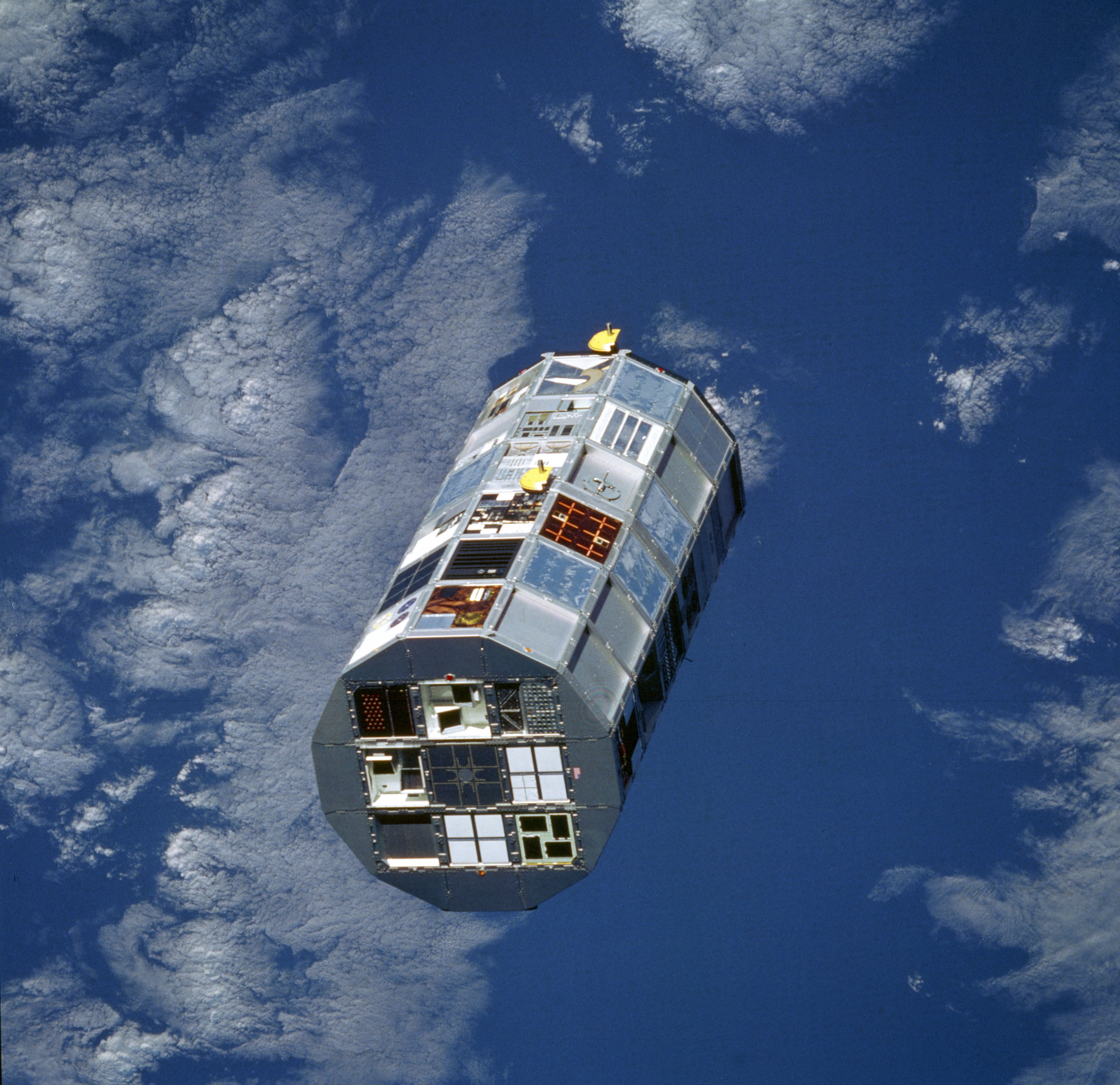 A cylindrical satellite covered in squares of different colors and materials floats in orbit above Earth
