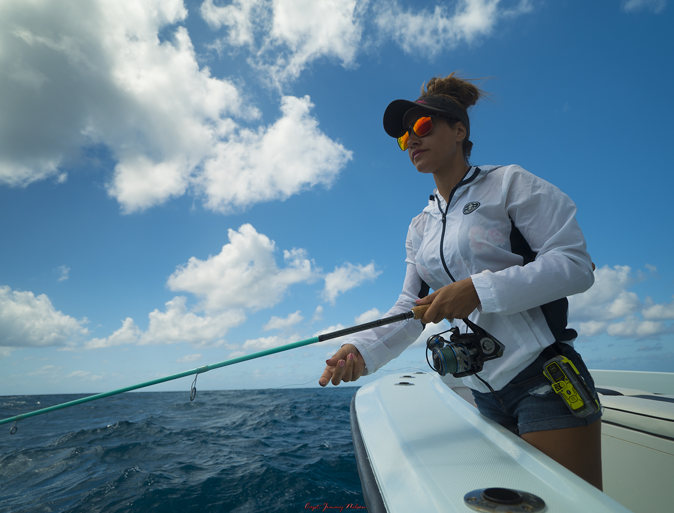 A woman on a boat with a fishing pole. Water, blue sky, puffy clouds in the background. 