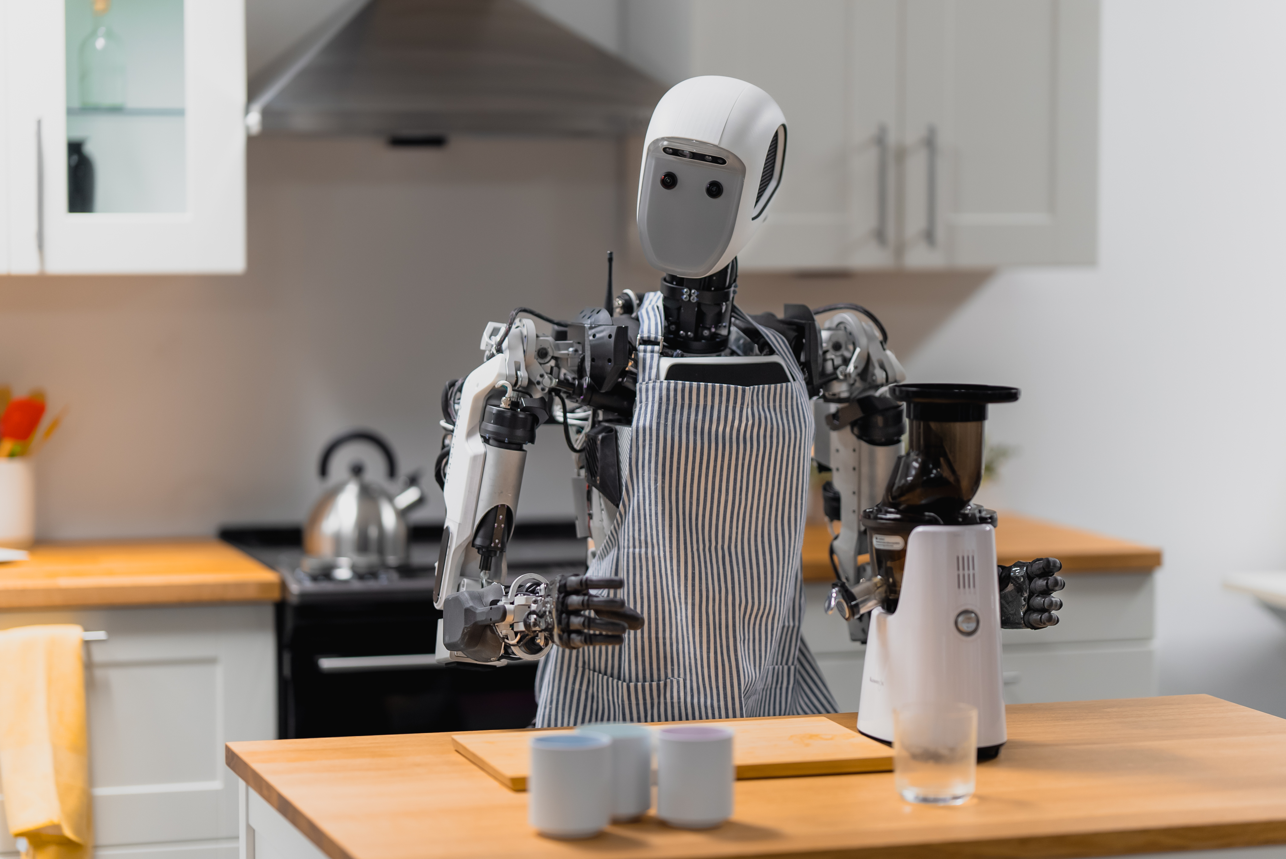 A humanoid robot wearing an apron in a kitchen setting, standing at an island with a juicing machine. 