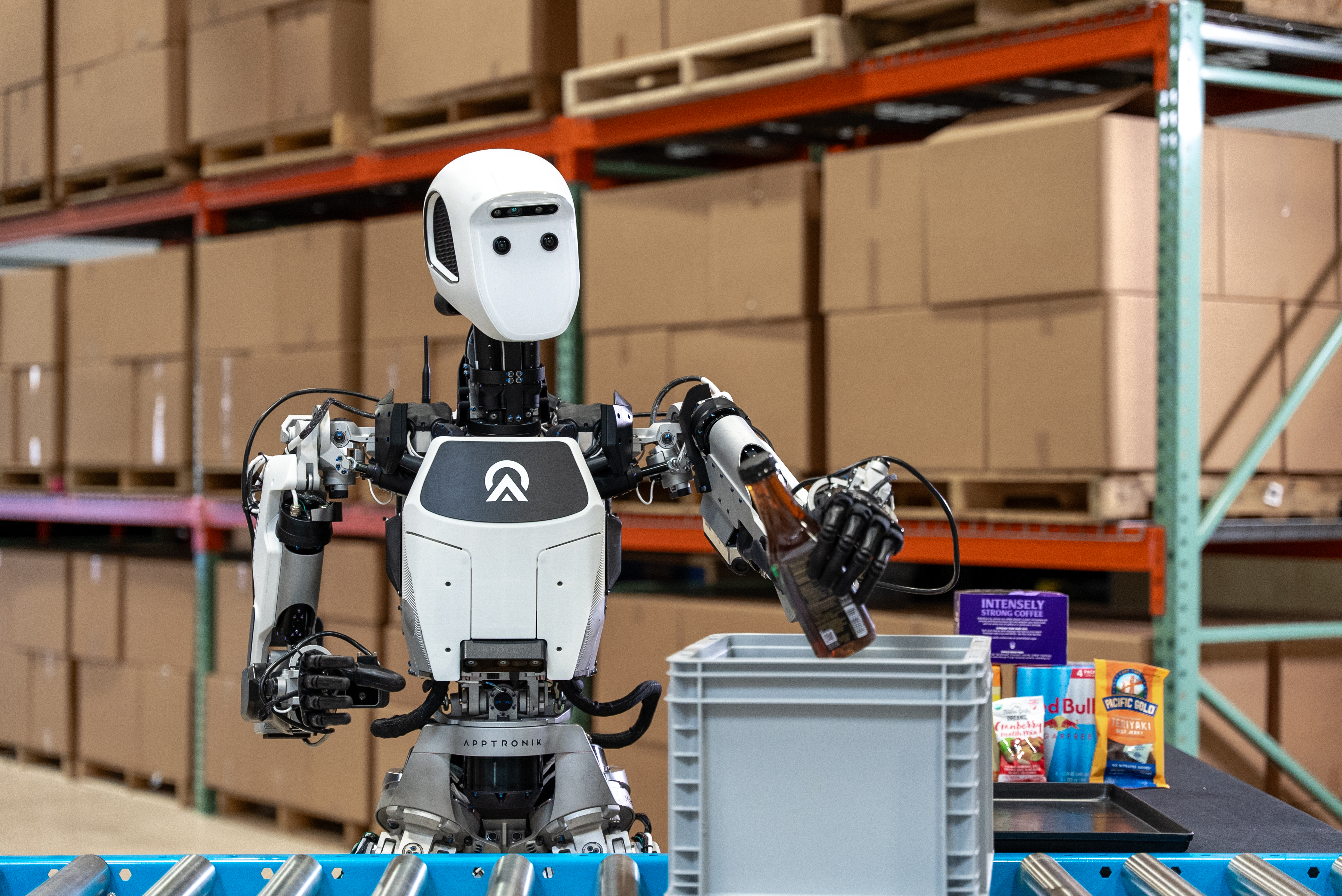A humanoid robot puts an amber glass bottle in a crate as it passes by on a factory assembly conveyor system. 