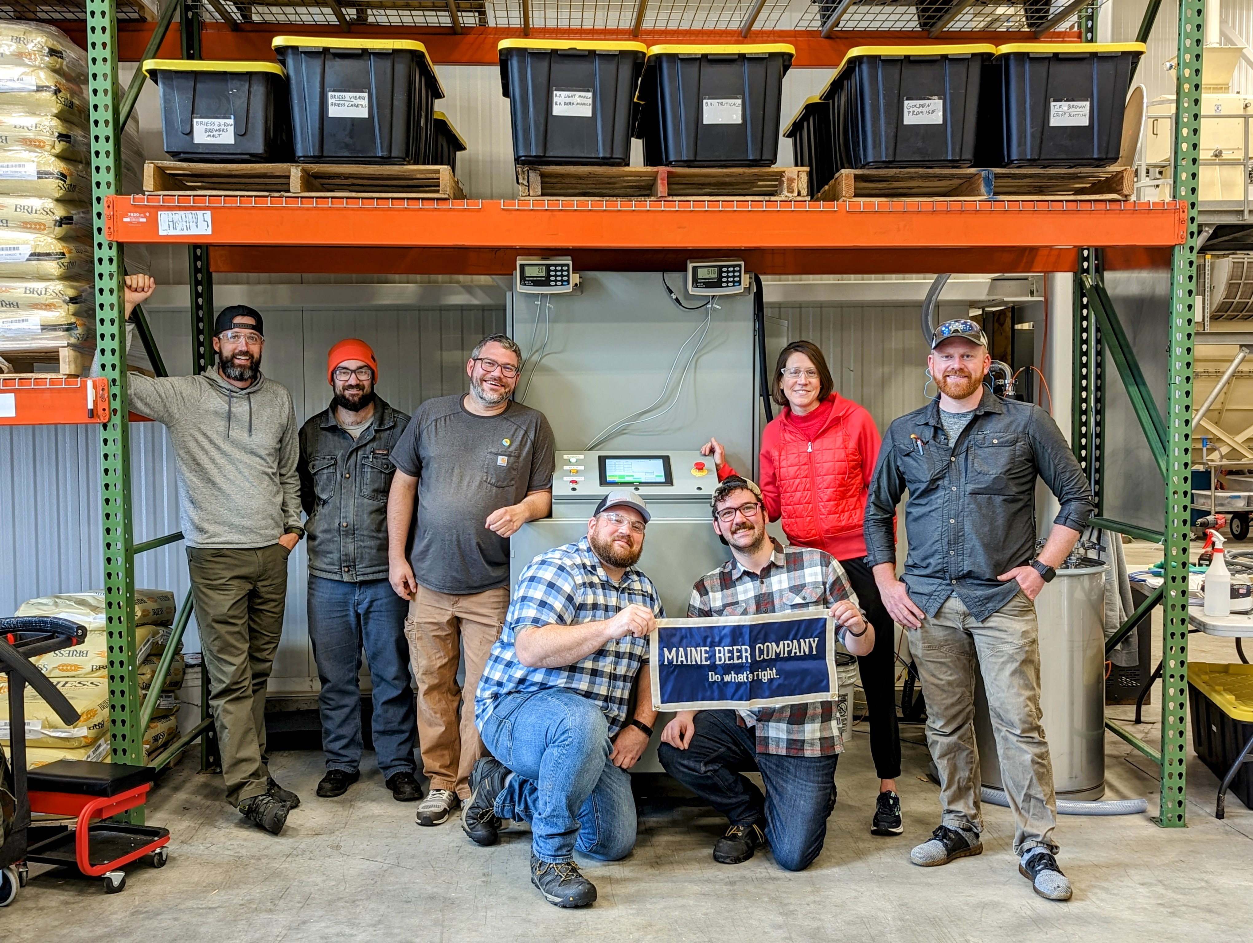 Seven smiling people stand around a machine, with two in the front kneeling and holding a sign that says “Maine Beer Company.” 