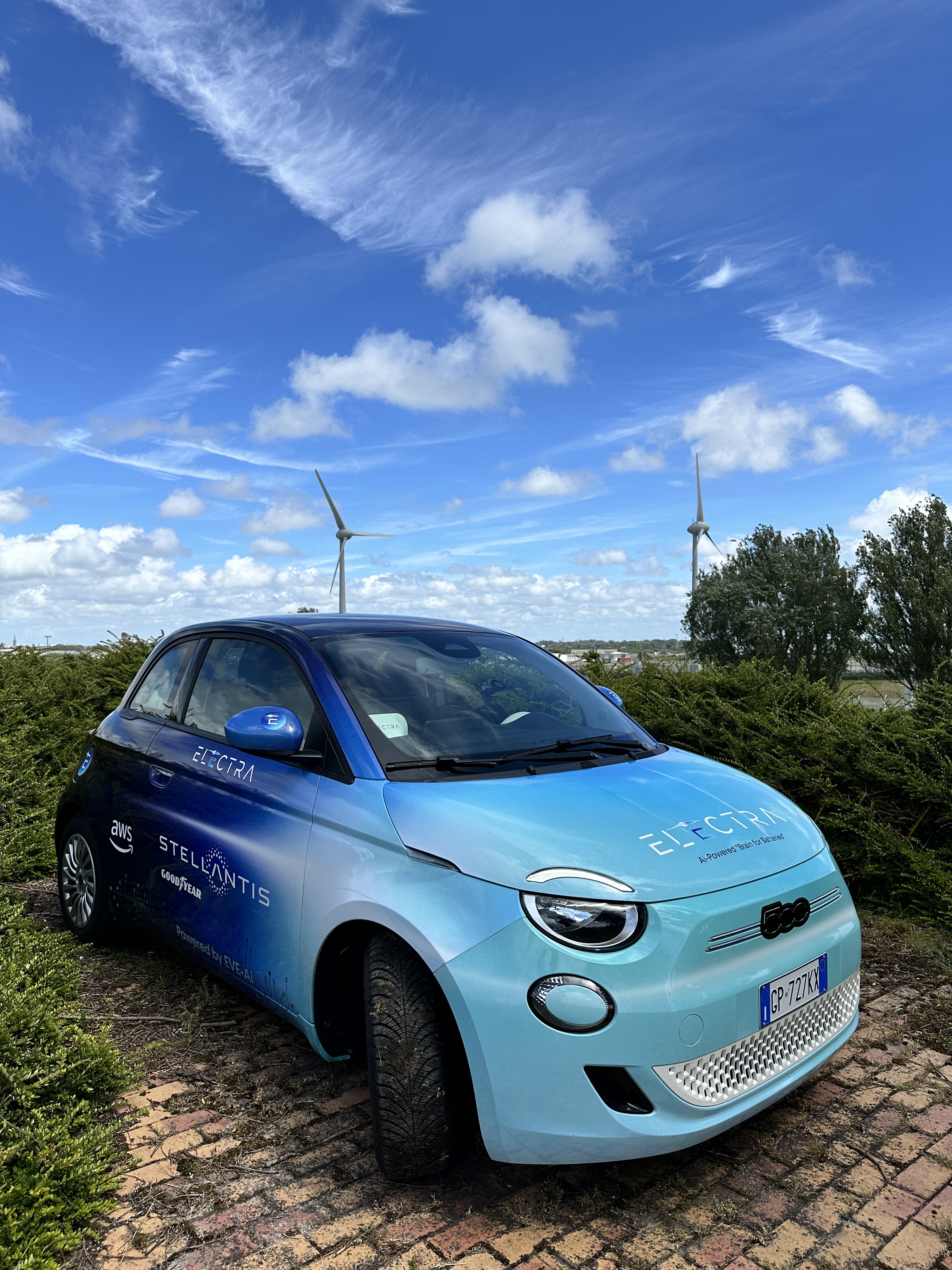A blue compact car covered in decals advertising Electra and the company’s partners is parked on a brick road. In the background are trees, bushes, and wind turbines.
