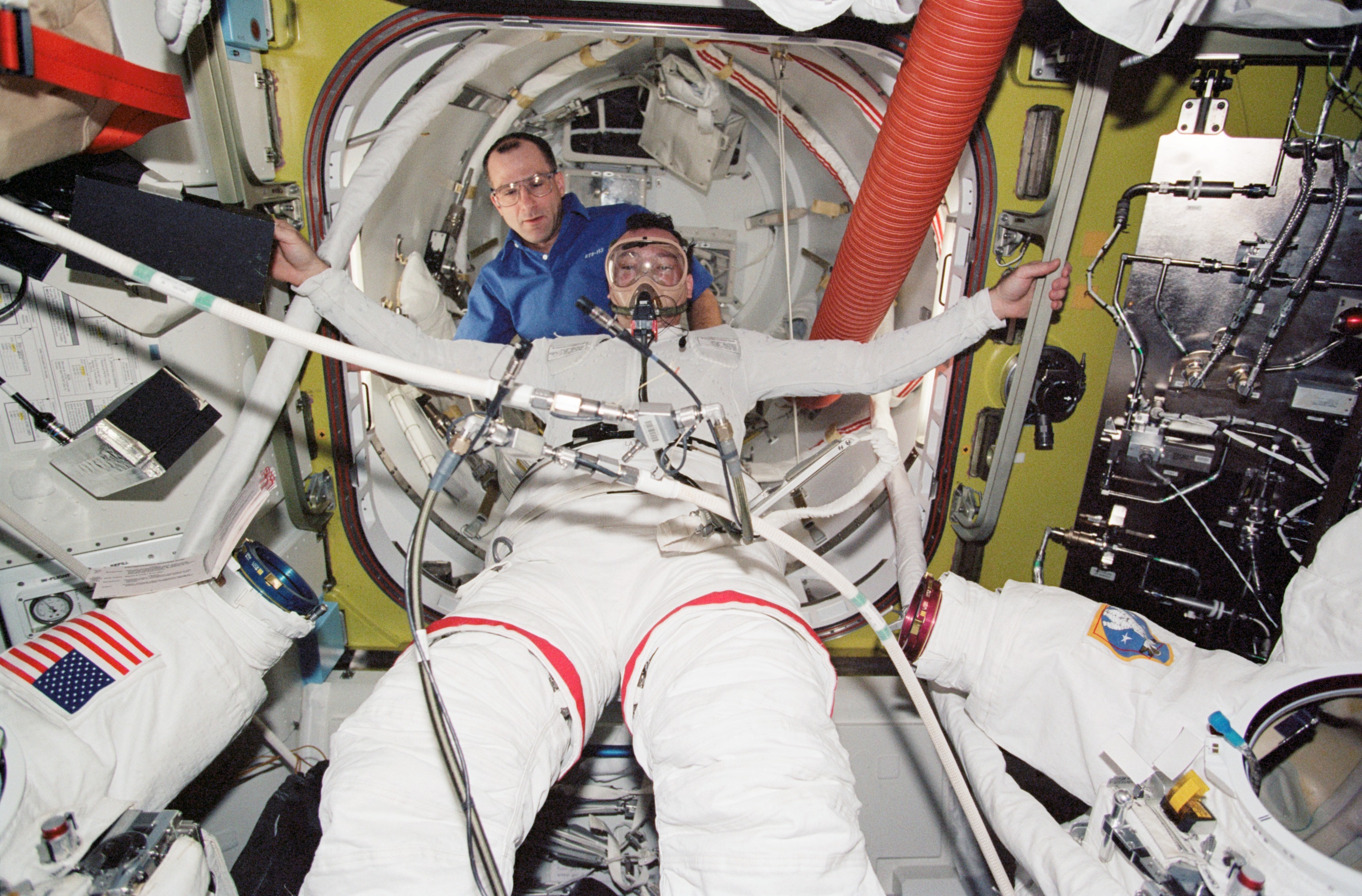 Astronaut inside a spacecraft undergoes a medical exercise with breathing apparatus while another crew member assists.