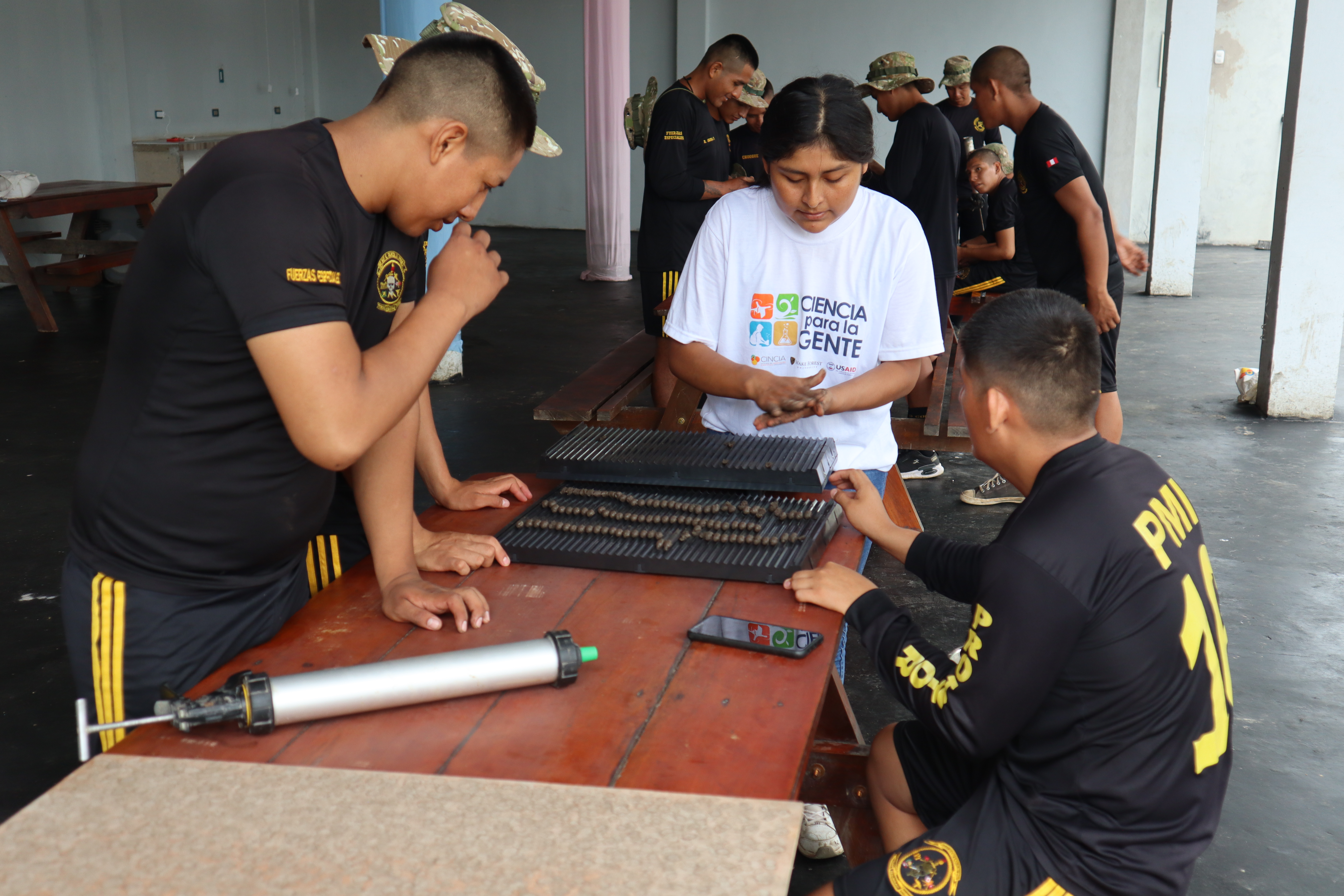 A woman hand molds soil-seed balls over a table with a collection of the balls as several men wearing black uniforms watch. Other people are gathered in the background. 