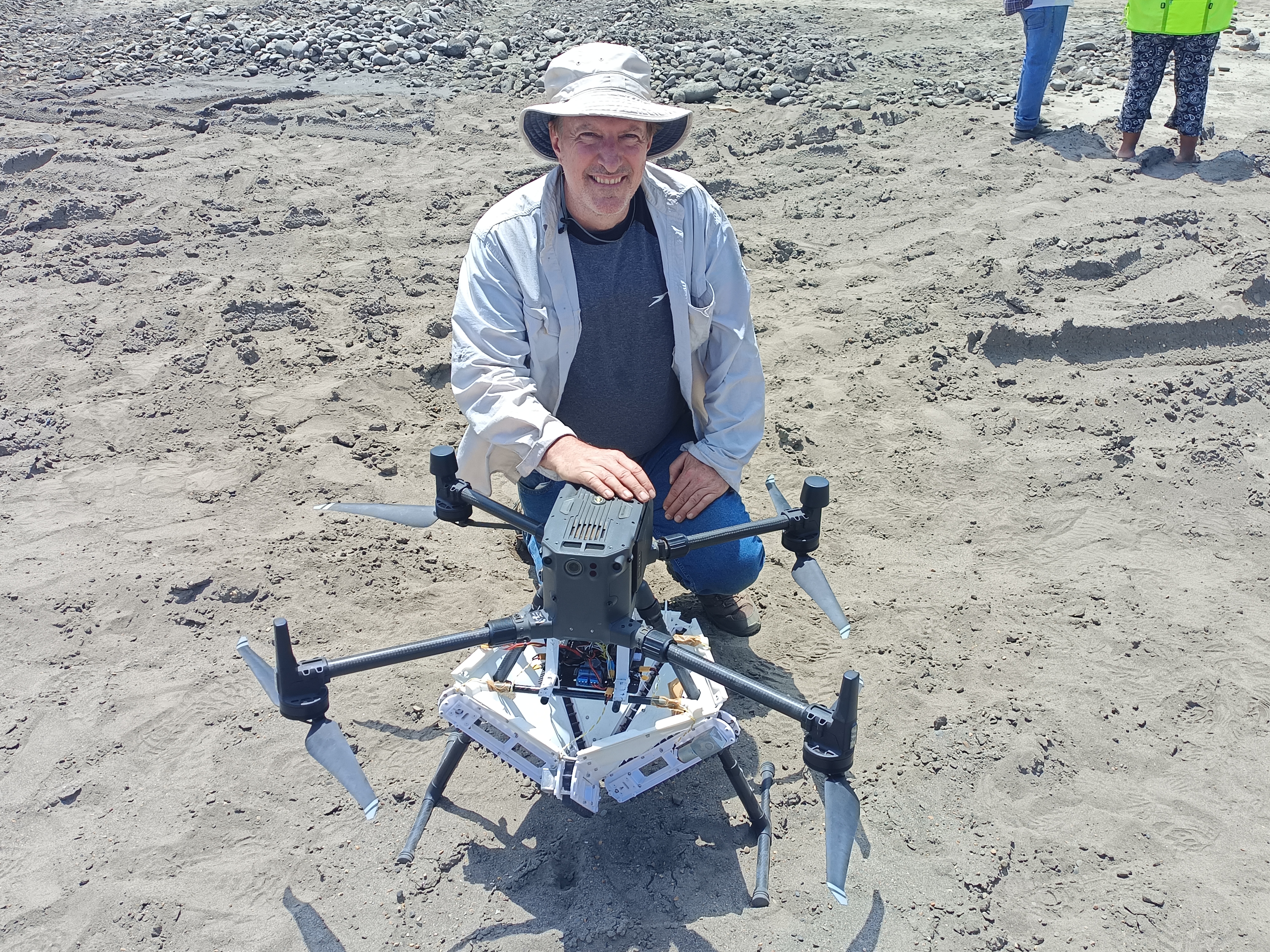 A man crouches next to a drone on land covered in gray sand or silt.  