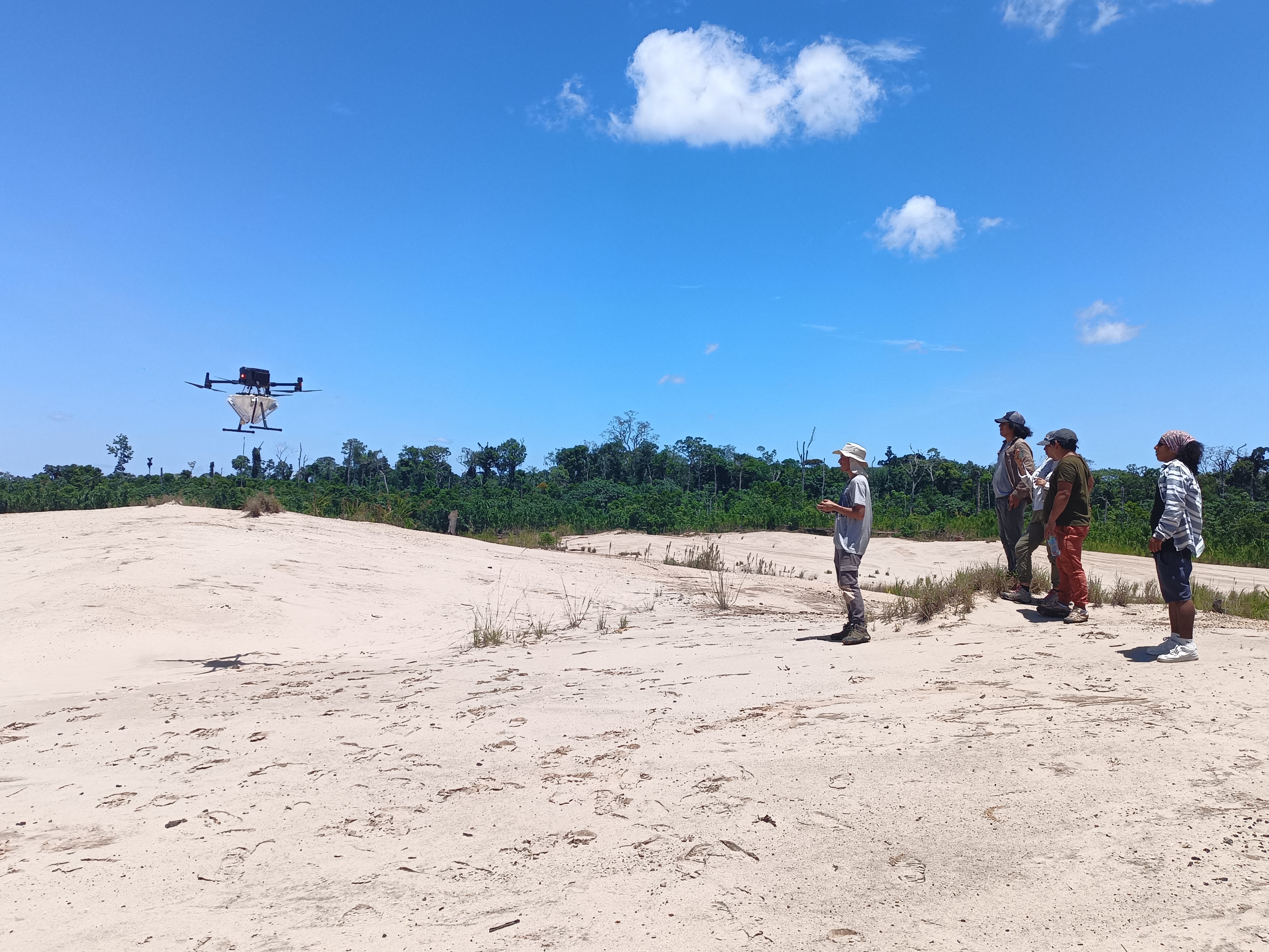A man remotely controls a drone with cargo as several people look on. 