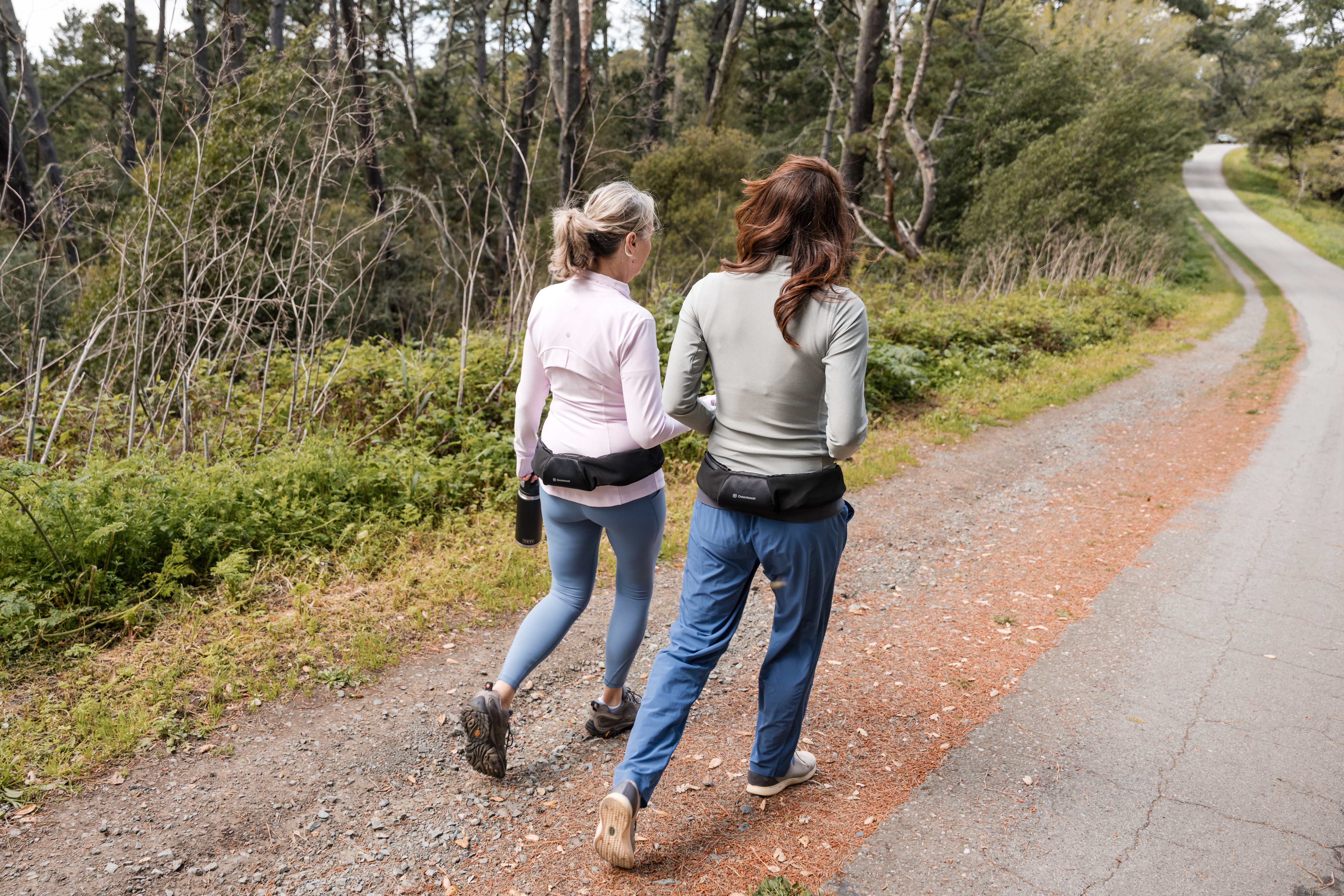 Two women walking along a trail in autumn, they are wearing Osteoboost Health belts around their waists