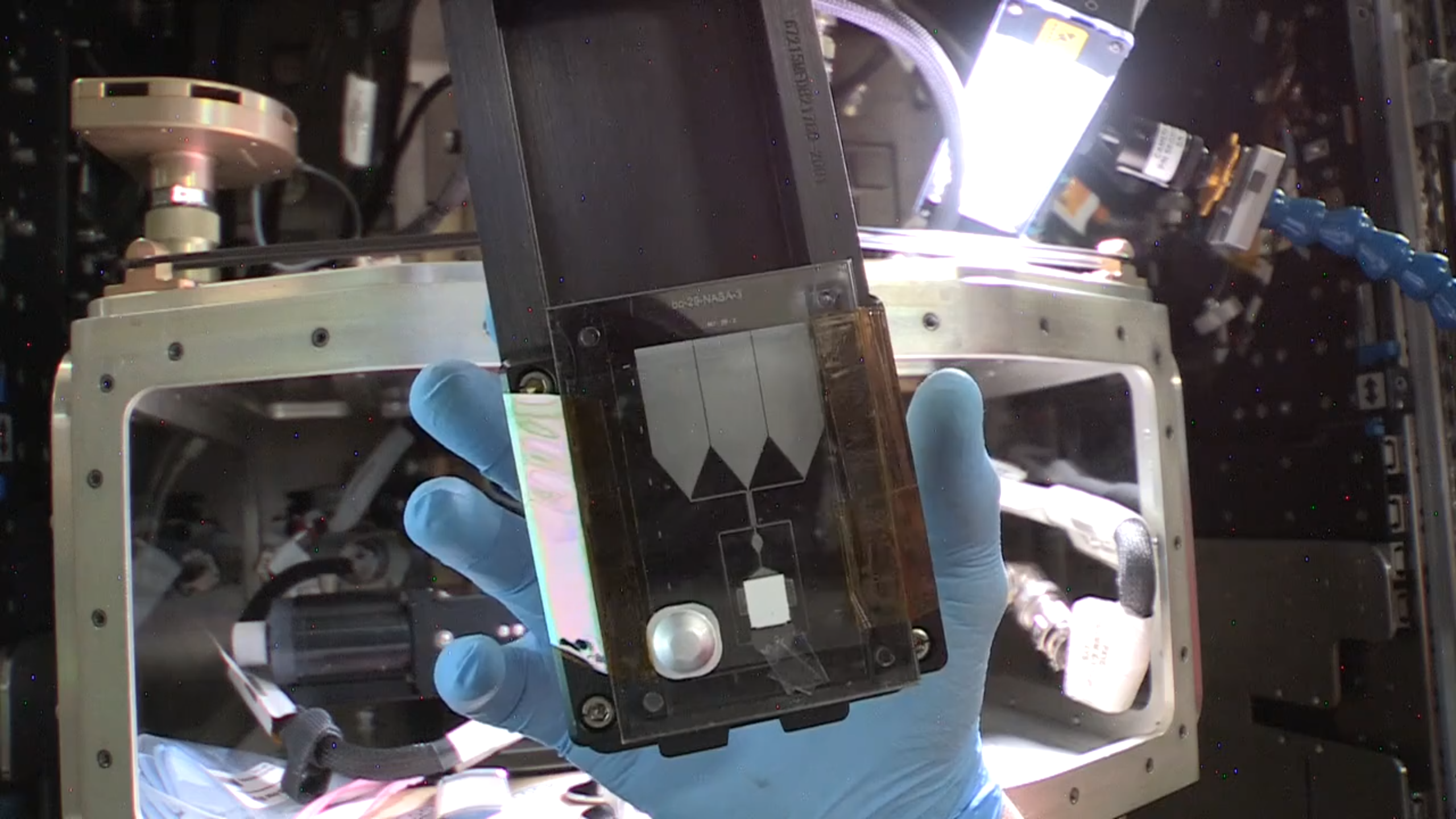 A gloved hand holds a device. On it, three triangles point down towards a white box.