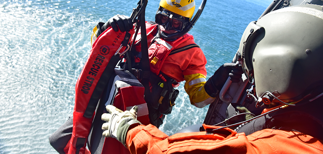 Rescue worker in orange gear hoists a person in a rescue strop from the ocean into a vehicle, likely a helicopter. 