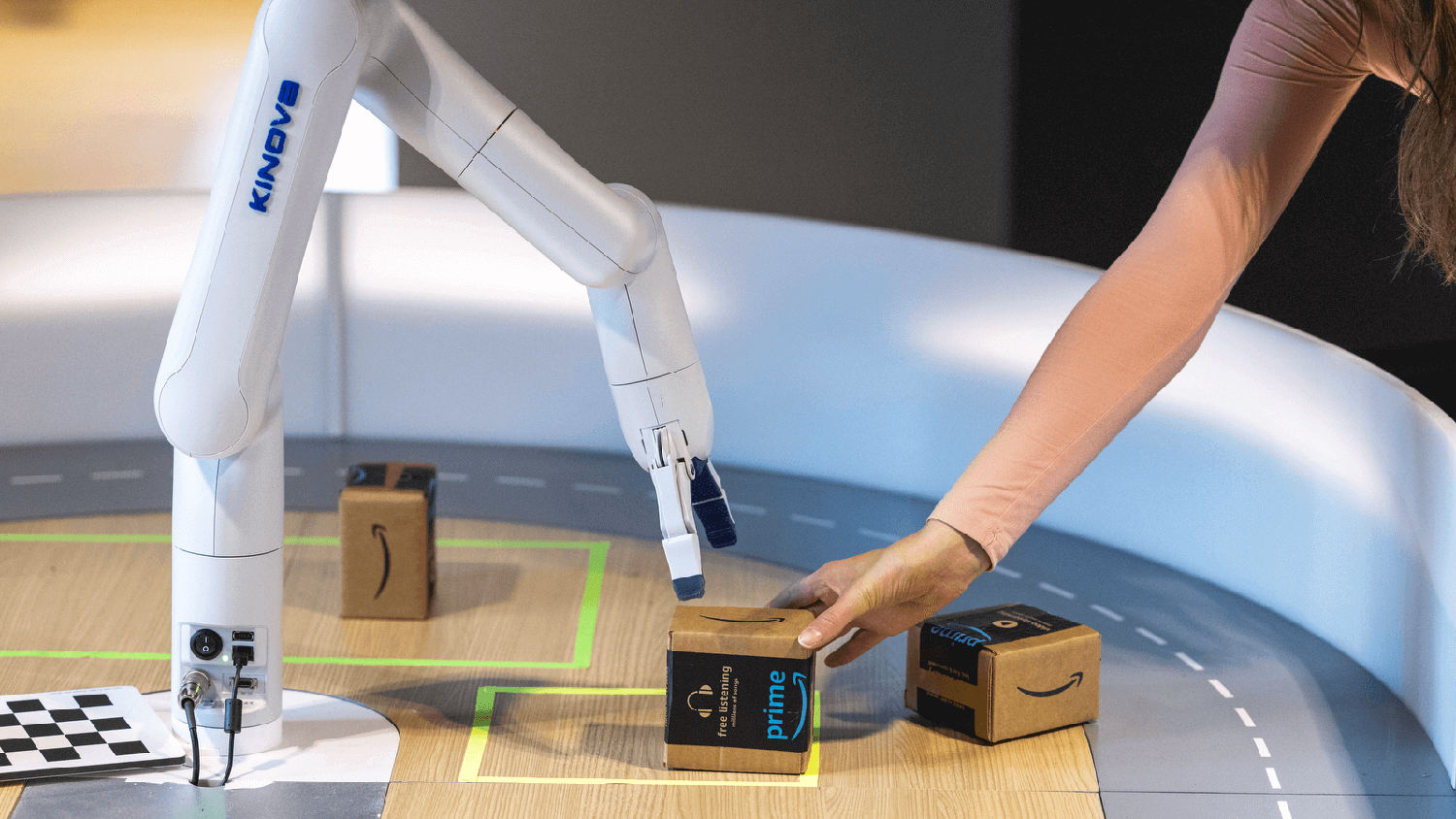 On a wooden table, a robotic arm reaches to pick up a small cardboard box being placed down by a human hand
