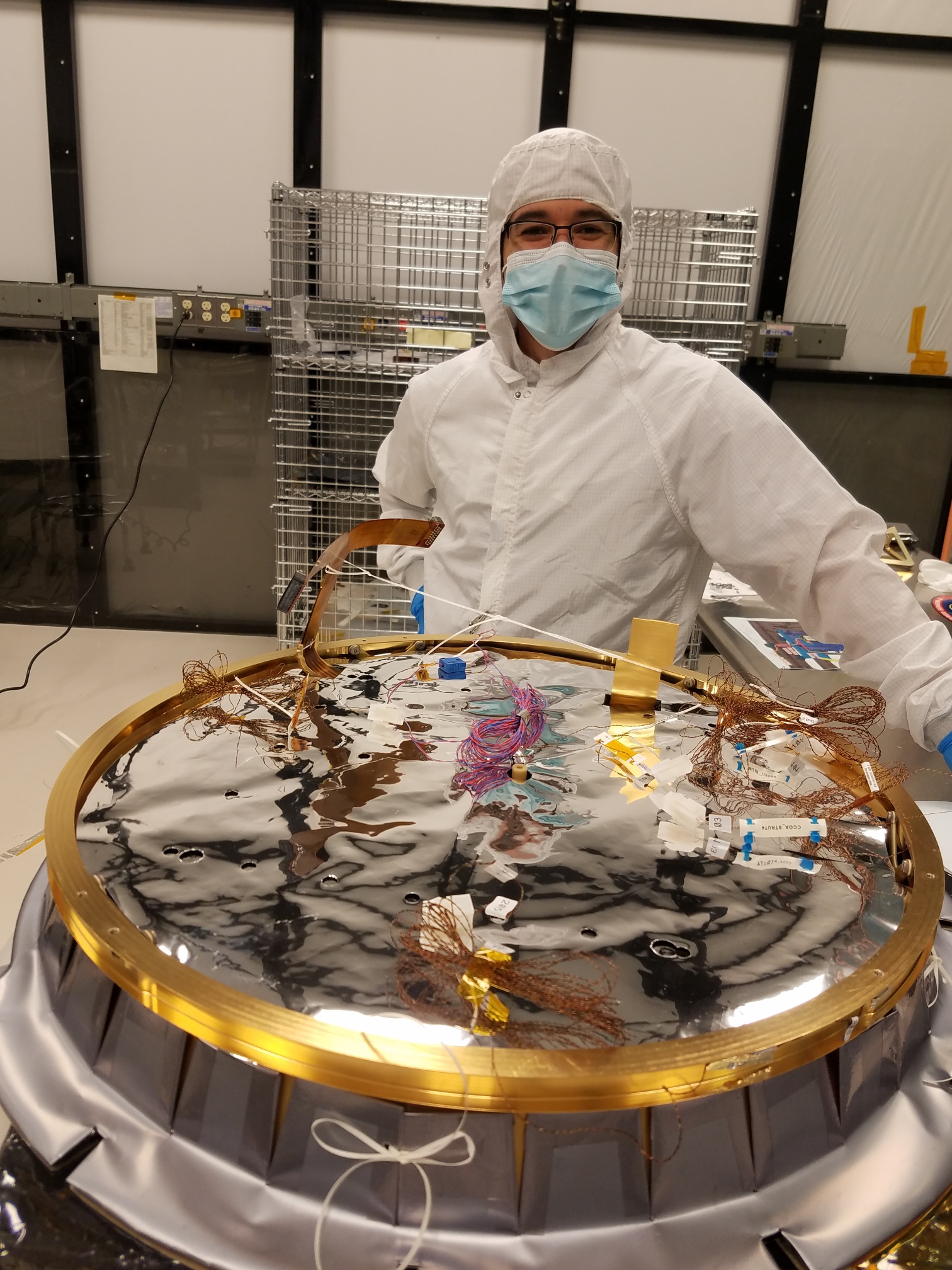 Technician stands wearing a white protective suit beside a circular piece of a spacecraft in-process
