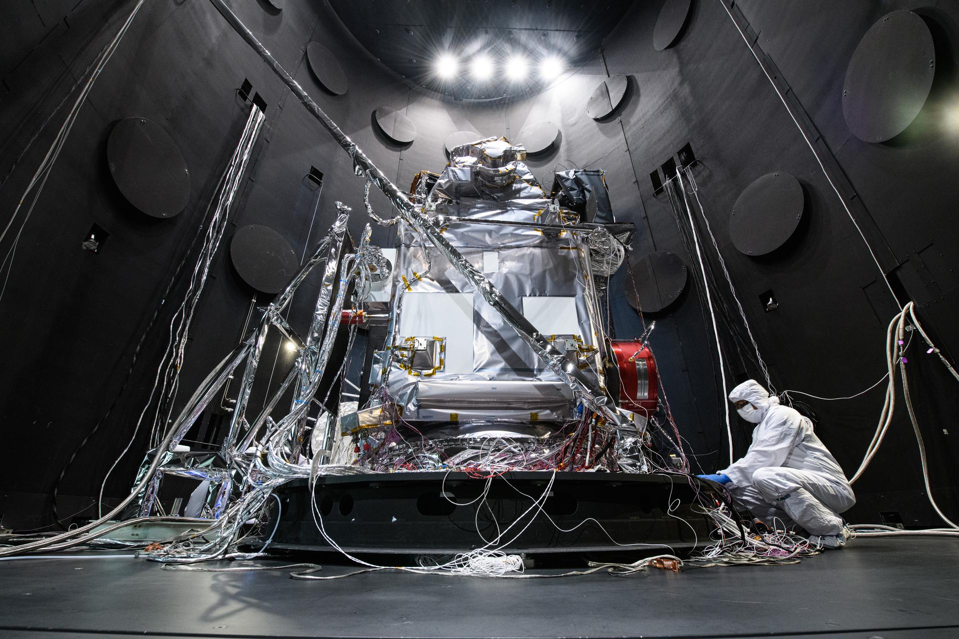 In a dark chamber, a spacecraft sits on a platform while an engineer in a clean room suit works on it