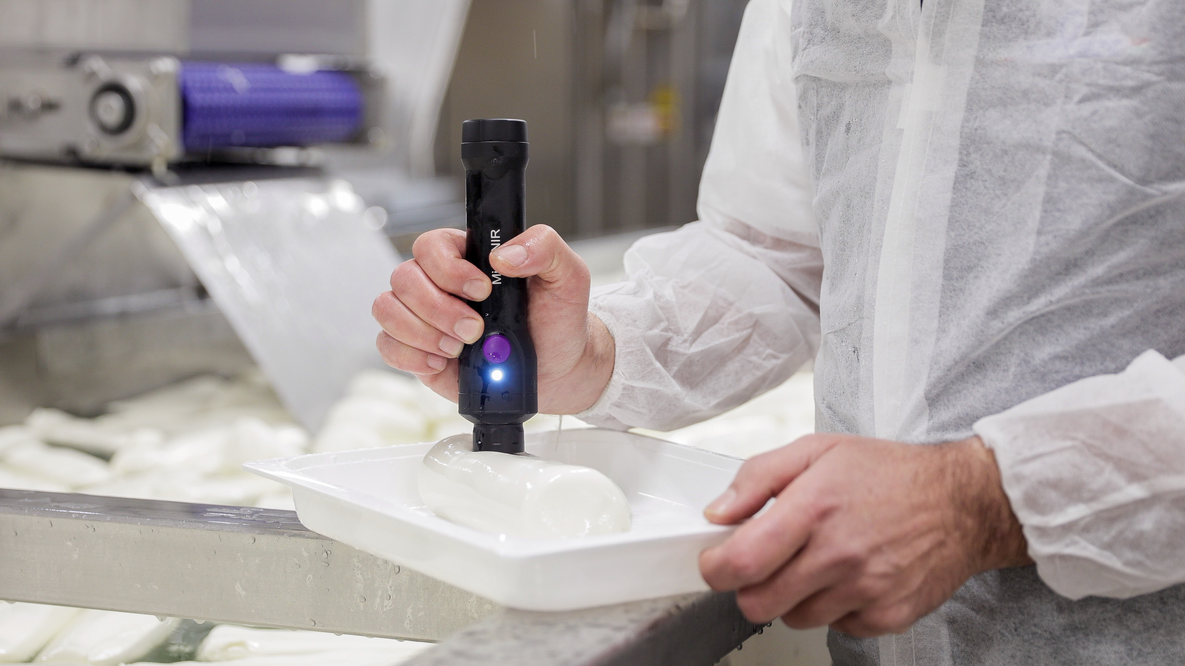 Close-up of a man’s hand holding a MicroNIR-branded device that is pressing against a white gelatinous substance in the shape of a cylinder in a tray