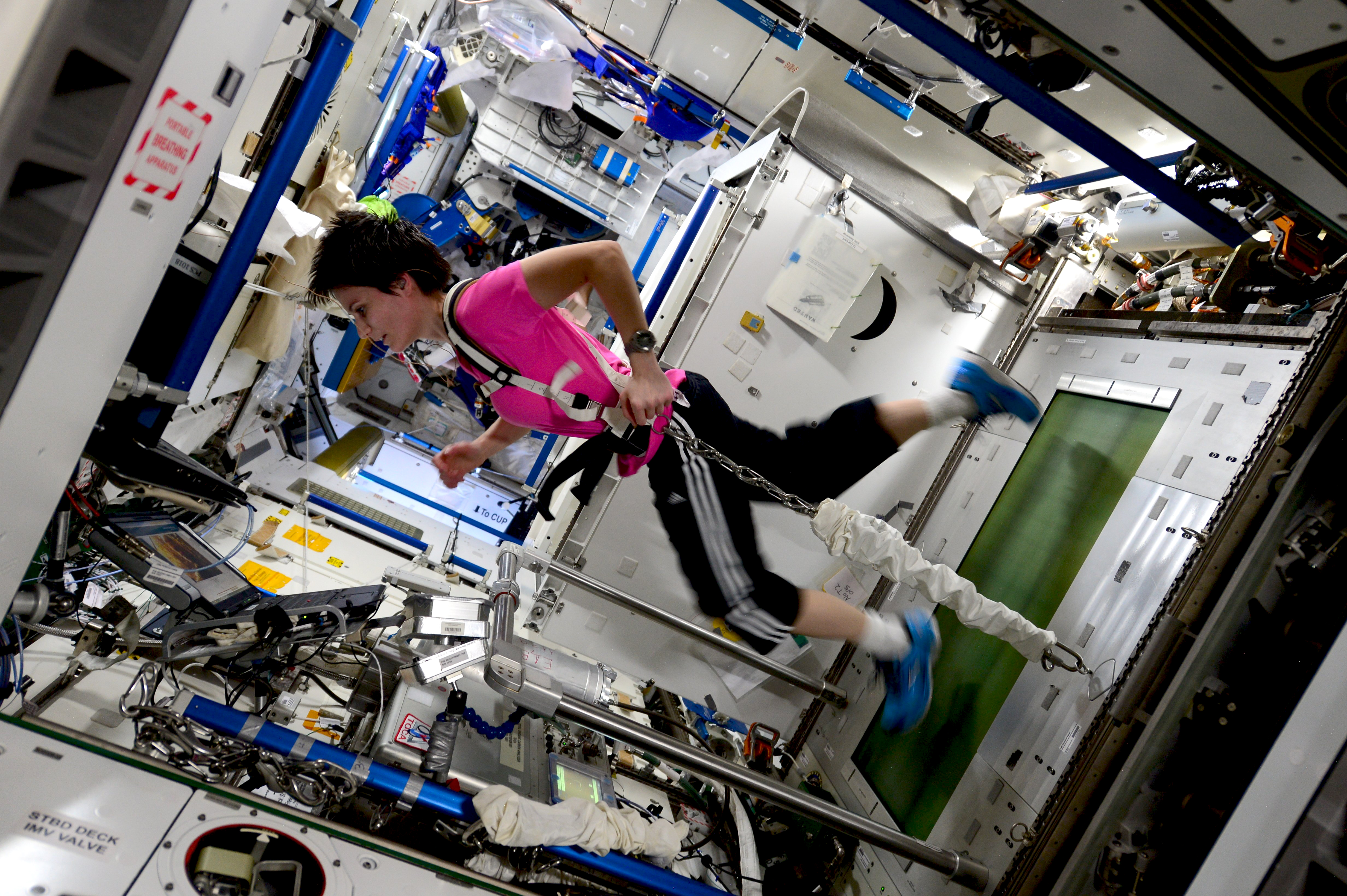 An astronaut running on a treadmill aboard the International Space Station, she is connected to the treadmill via an elastic harness.