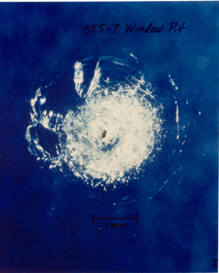 On a blue background, cracks of glass radiate from a central point. Text written on the image in black marker reads “STS-7 Window Pit.”