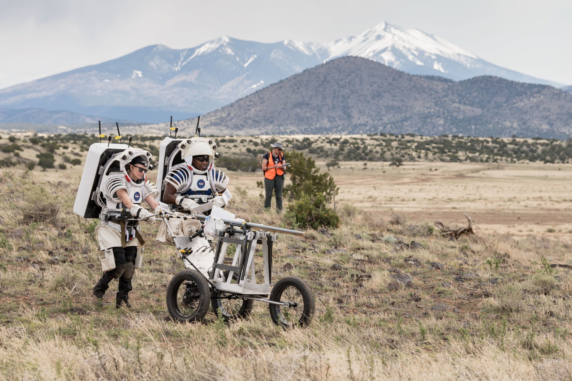 In a mountainous environment, Two people wearing mock spacesuits push a cart while another person in a high visibility vest writes on a clipboard in the background.