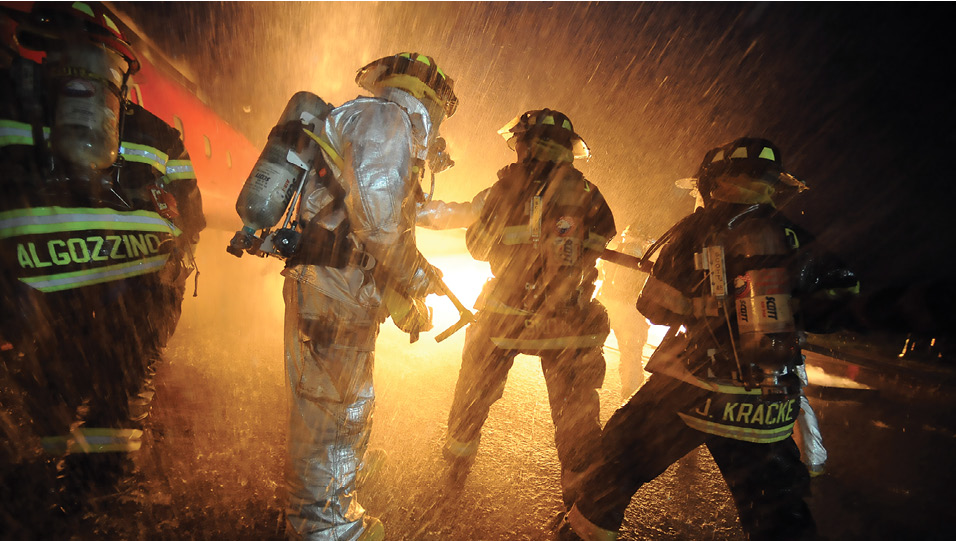Four firefighters wearing oxygen tanks hose down a blaze in the rain