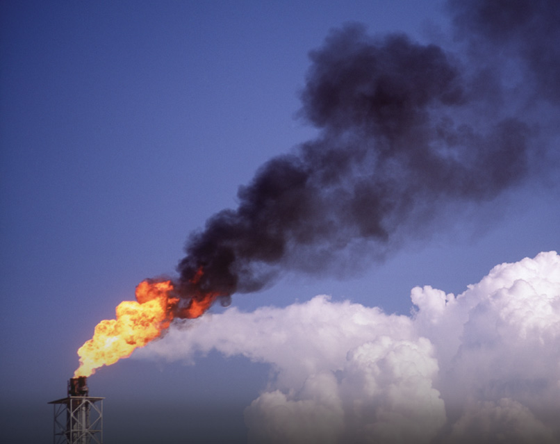Flames and smoke flare from the top of an oil well into the blue sky