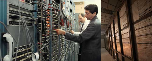 Two men plug and unplug wires attached to a wall of ports. Digital system covers the wall with no visible cables