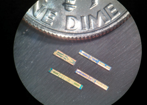 A grouping of four Freedom Photonics laser chips, shown with a dime for scale. Each is around the size of a letter stamped on the dime