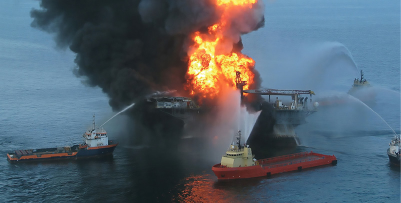 Barges spray water on large fire burning on an offshore oil well with black smoke billowing out