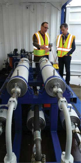 Two men with reflective safety vests standing near a stack of cylindrical SureShear chambers