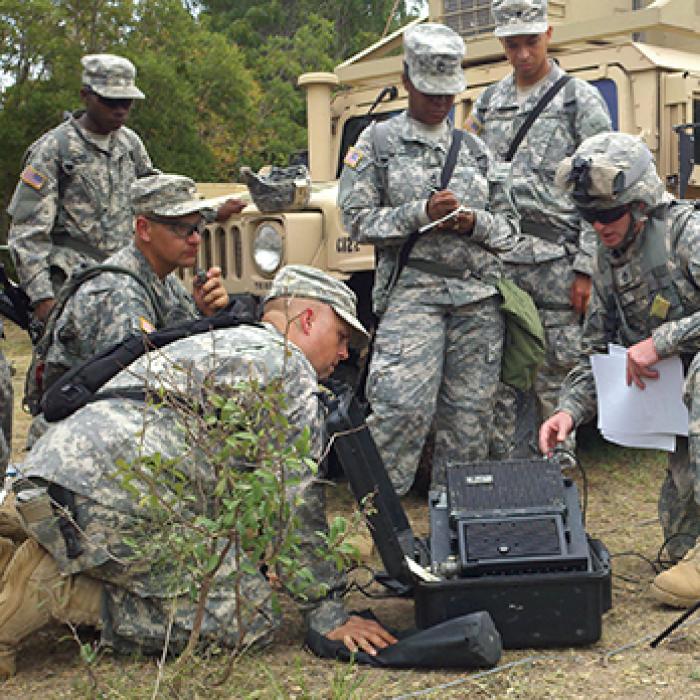 Service members in camouflage dress crowd around field communication equipment