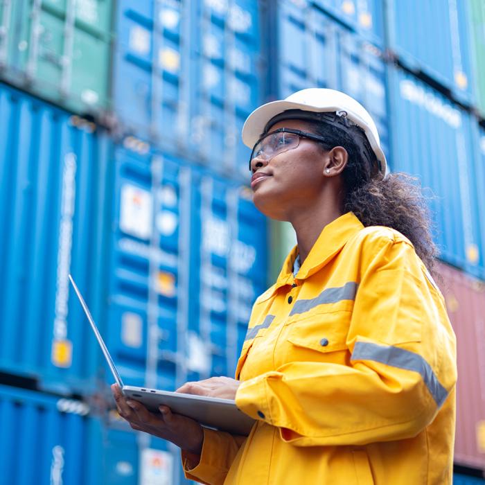 A woman holds a laptop in front of stacked shipping containers