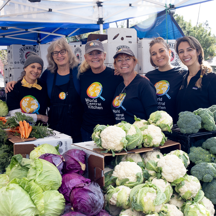 Women wearing World Central Kitchen T-shirts stand behind an assortment of vegetables 