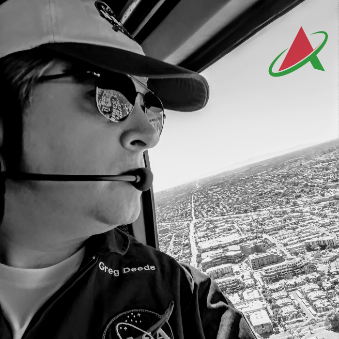 A black and white photo of a man wearing a shirt emblazoned with the NASA emblem looking out of the window of a helicopter at the city below.