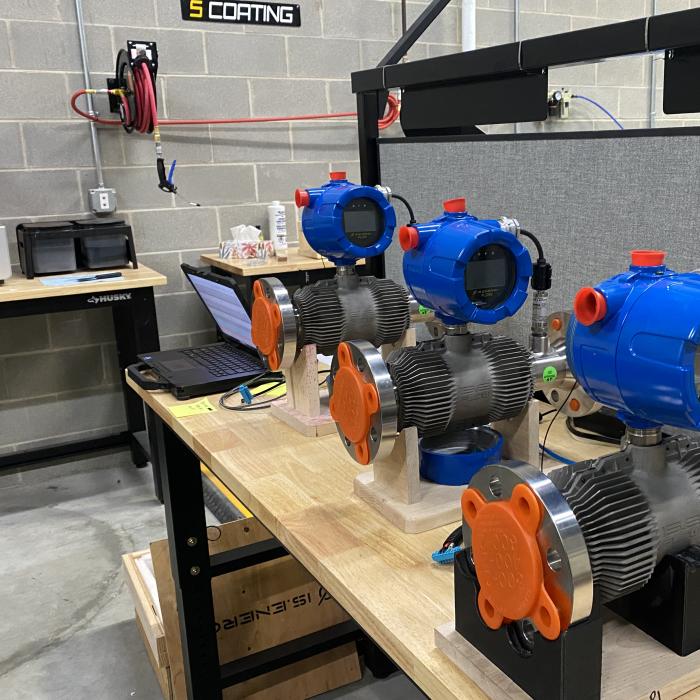 On a wooden table are three blue and orange machines next to a laptop computer