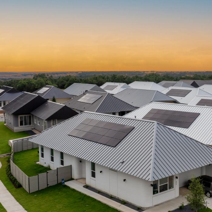 Twenty-some closely set houses with similar roofs, designs, and yards. They are part of a suburban complex, with some trees in the background. 