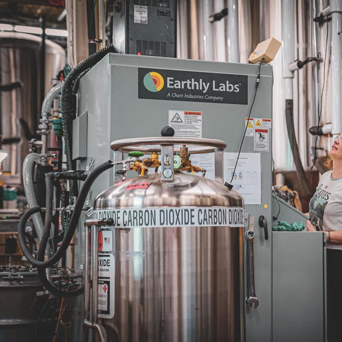 A woman controls an Earthly Labs-branded machine in a factory setting; a tank that says “carbon dioxide” is in the foreground. 