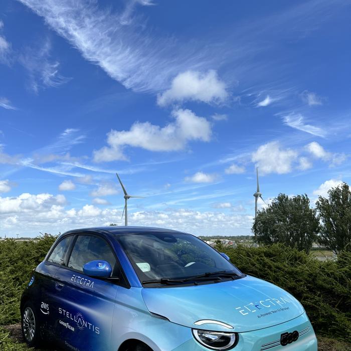 A blue compact car covered in decals advertising Electra and the company’s partners is parked on a brick road. In the background are trees, bushes, and wind turbines.